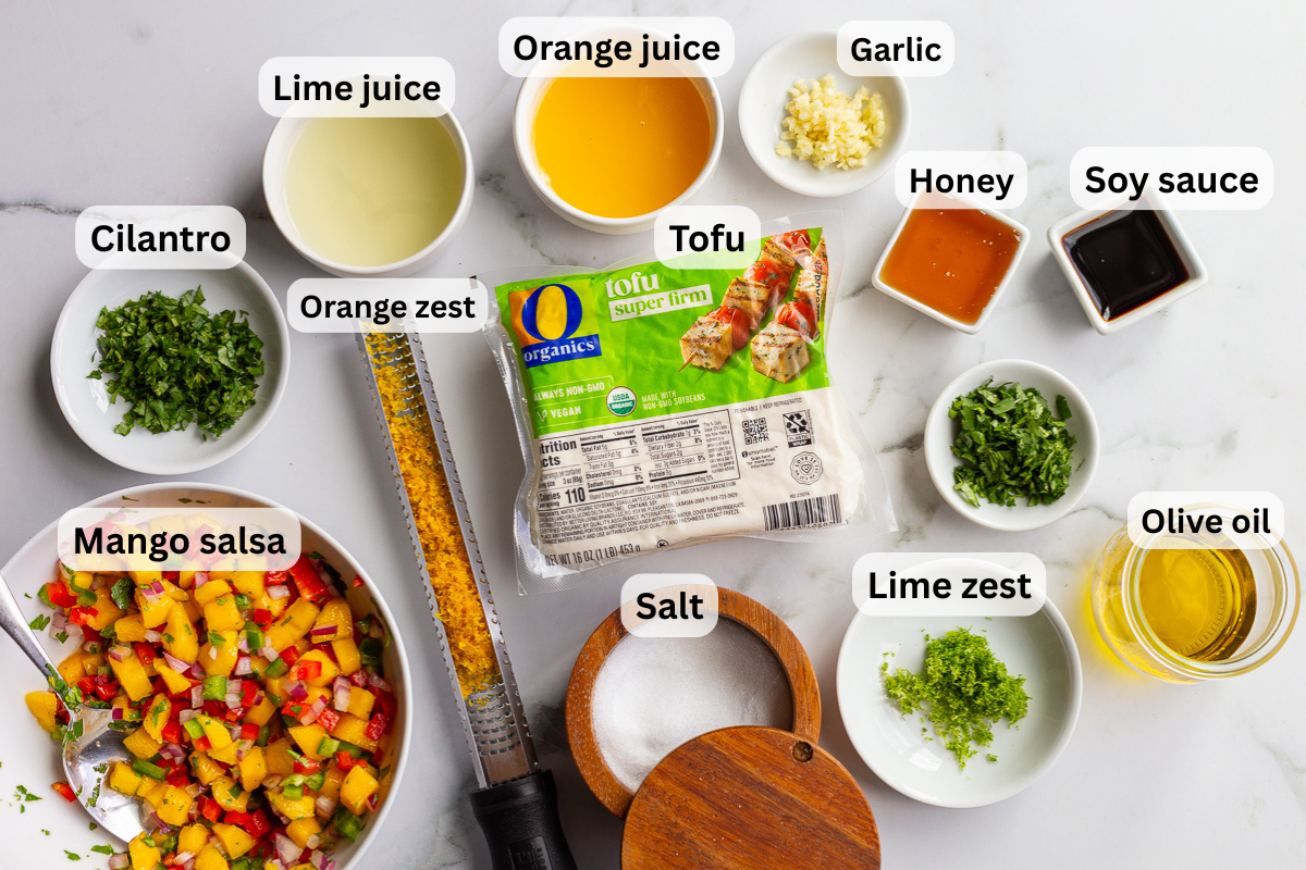 Overhead shot of cilantro lime tofu ingredients including super firm tofu, lime juice, lime zest, orange juice, orange zest, garlic, honey, soy sauce, cilantro, olive oil, salt, and a bowl of mango salsa, each labeled on a marble countertop.