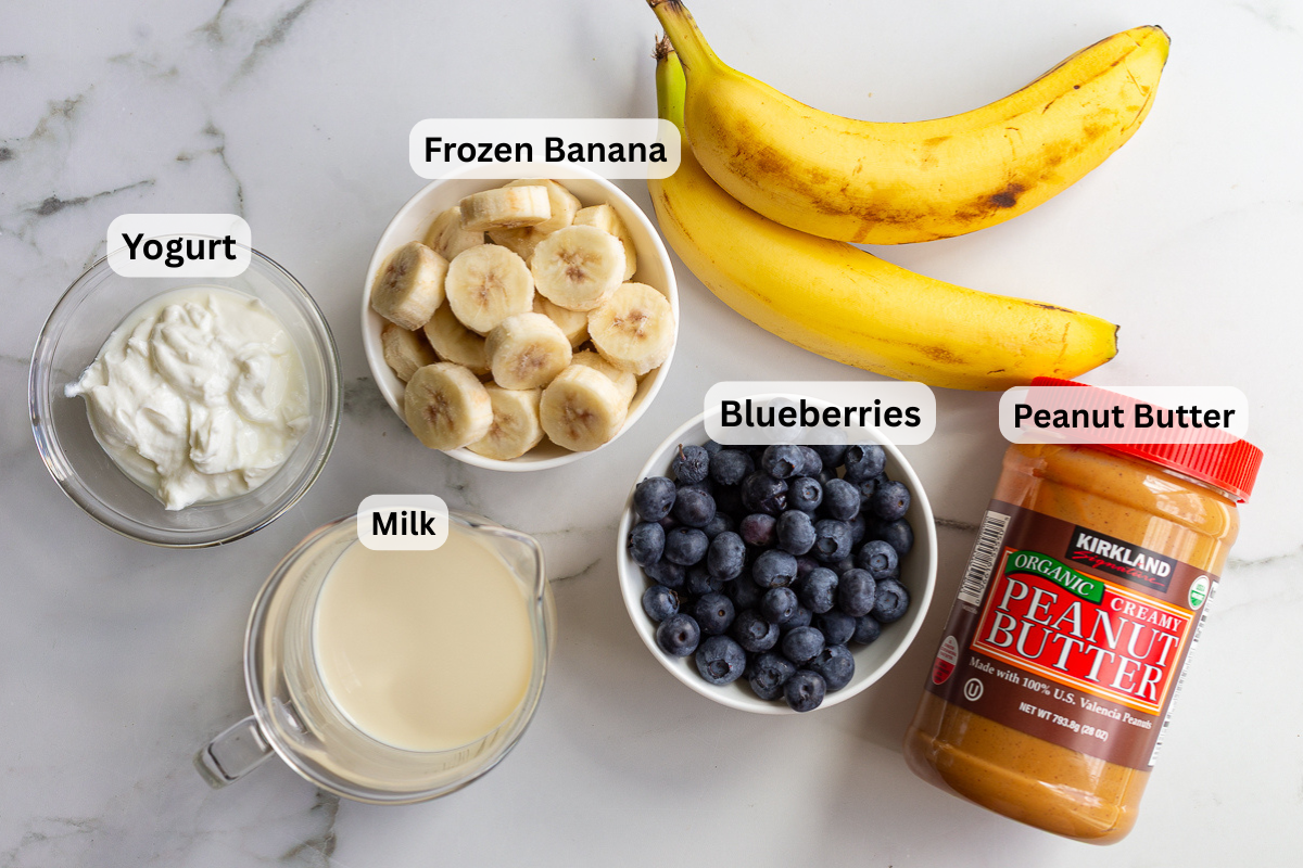 Overhead shot of yogurt, frozen banana, blueberries, milk, and peanut butter labeled and arranged on a marble counter for a blueberry smoothie