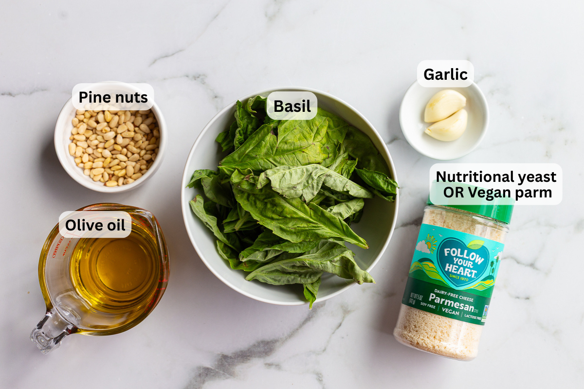 overhead shot of vegan pesto ingredients including fresh basil, pine nuts, garlic, and vegan parmesan on a marble counter