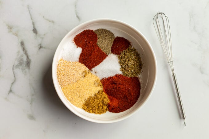 Individual portions of chili powder, smoked paprika, cumin, garlic powder, onion powder, cayenne, oregano, salt, and black pepper measured into a white bowl next to a small whisk on a white marble surface. used to make fajita seasoning