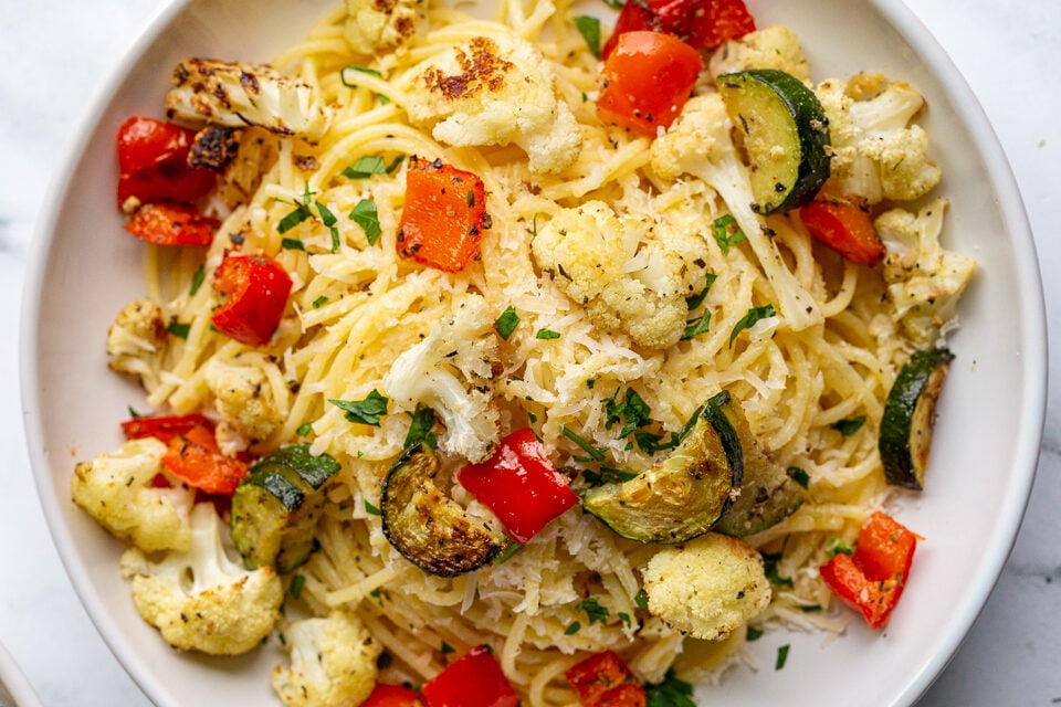 garlic butter noodles in a white bowl topped with parmesan cheese and parsley as well as roasted cauliflower, zucchini and bell pepper