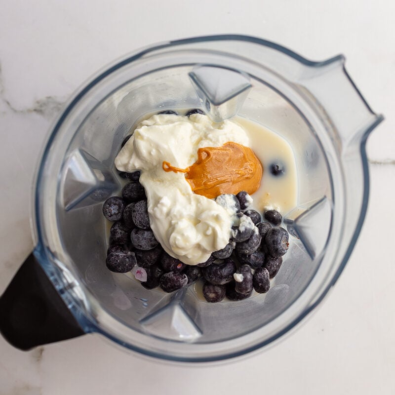 Overhead shot of a blender filled with frozen blueberries, banana, yogurt, and peanut butter before blending