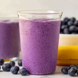 blueberry smoothie in a medium clear glass with blueberries and bananas in the background
