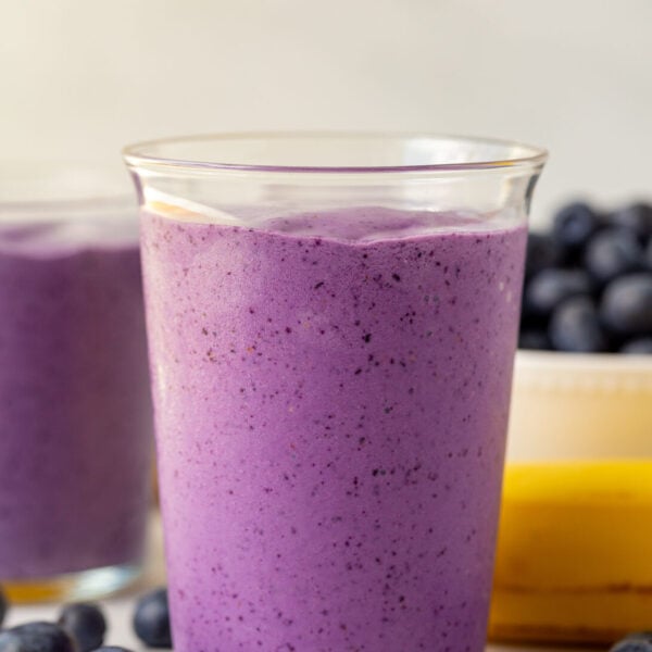 blueberry smoothie in a medium clear glass with blueberries and bananas in the background