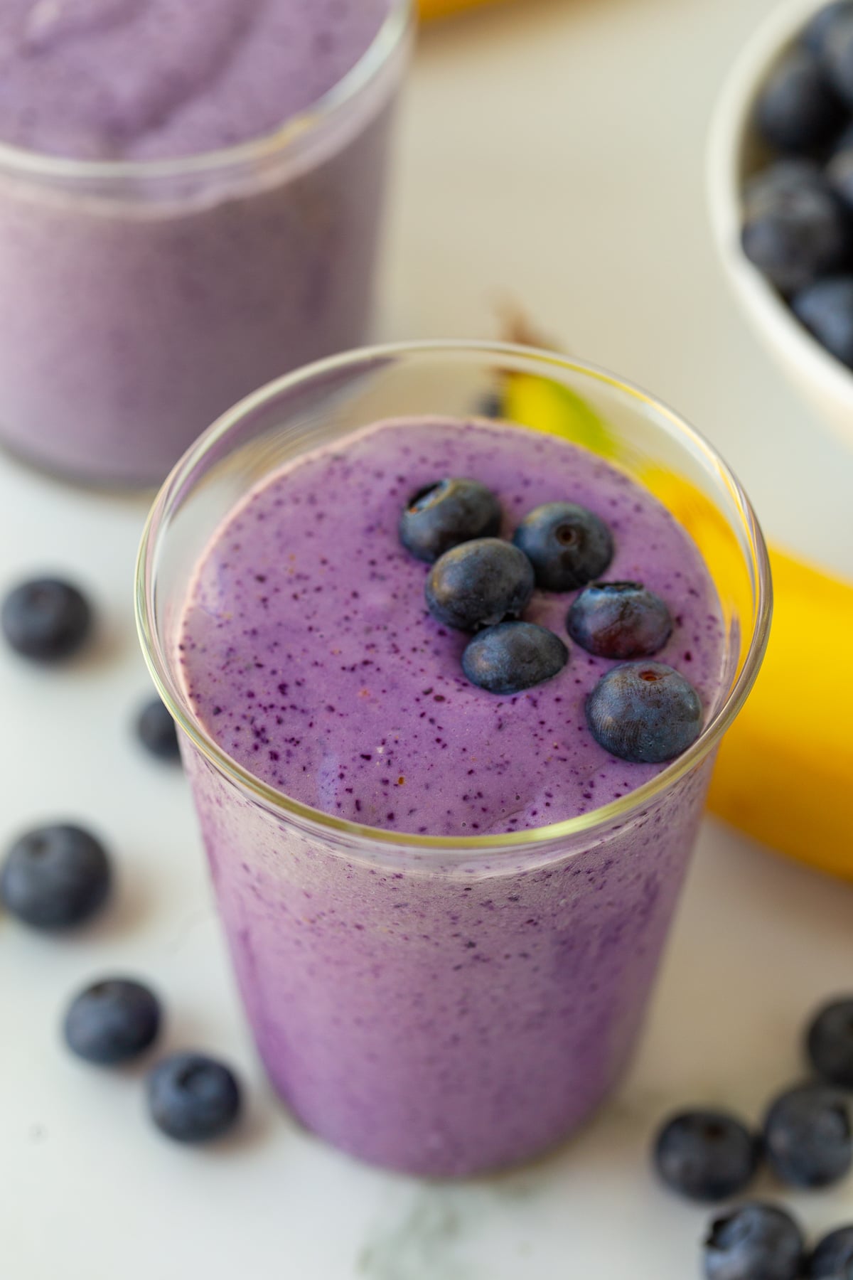 blueberry smoothie in a medium clear glass with blueberries and bananas in the background