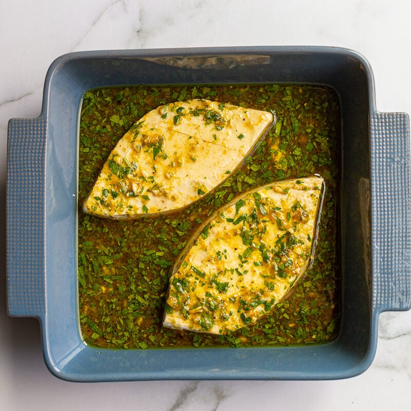 Two triangular slices of tofu marinating in a blue baking dish in a cilantro, lime, and garlic marinade.