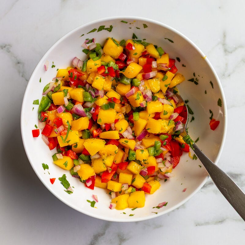 Overhead shot of mango salsa with diced mango, red bell pepper, red onion, and cilantro in a white bowl with a spoon.