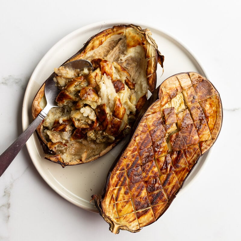 roasted eggplant halves on a plate, one scored and being scooped with a spoon