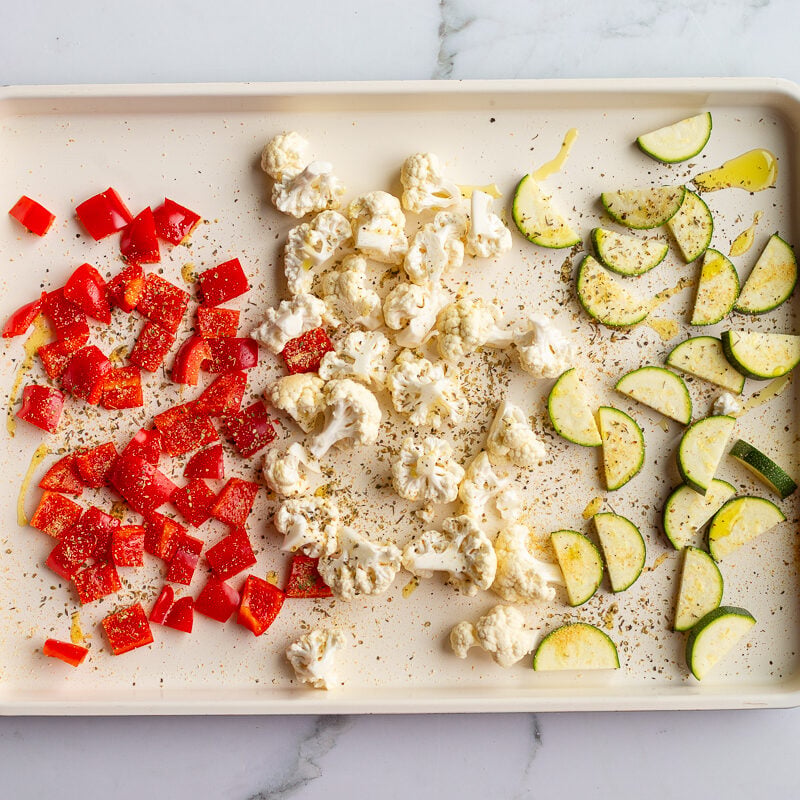 Diced red bell pepper, cauliflower florets, and sliced zucchini spread on a baking sheet and seasoned with olive oil, salt, pepper, and Italian seasoning, ready to go into the oven.