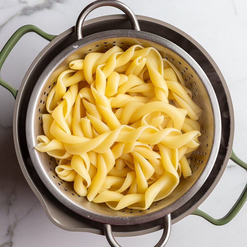 Cooked pasta shells draining in a metal colander on a marble countertop.