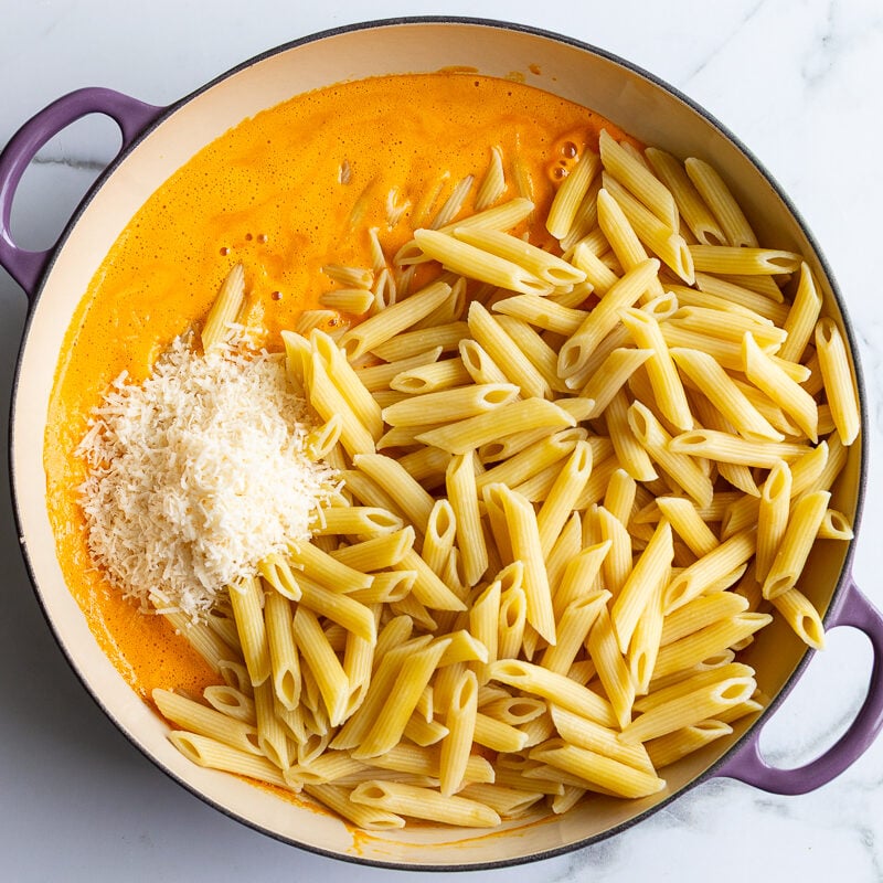 Cooked penne pasta and grated parmesan added to roasted red pepper sauce in a skillet