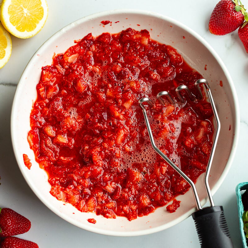 Mashed strawberries in a white bowl with a potato masher, lemon halves and fresh strawberries nearby