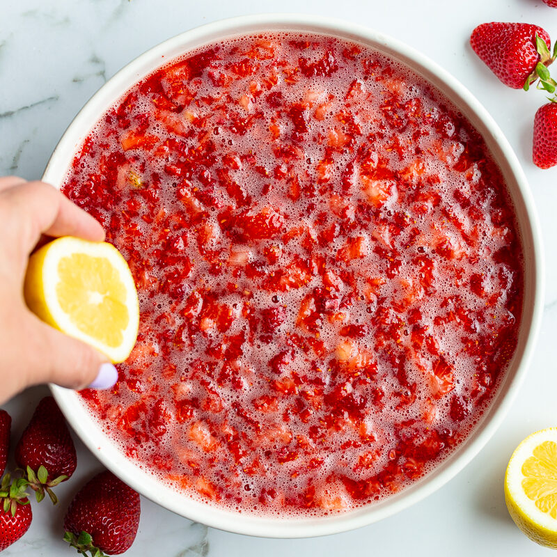 Hand squeezing lemon juice into a bowl of mashed strawberry jam mixture
