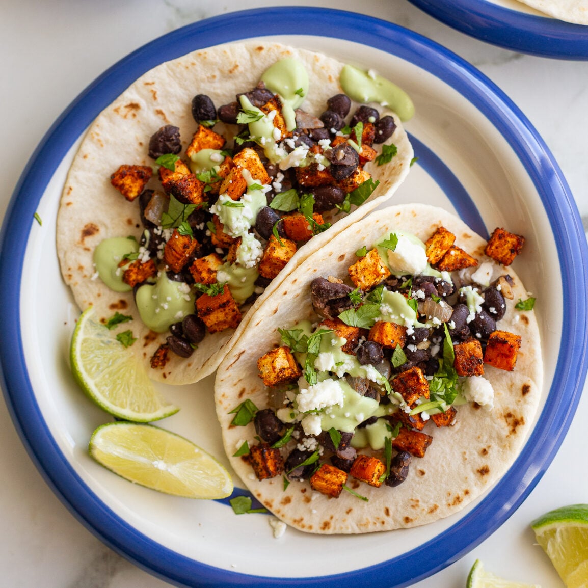 sweet potato and black bean tacos drizzled with an avocado crema. they're sitting on a white plate with royal blue rim