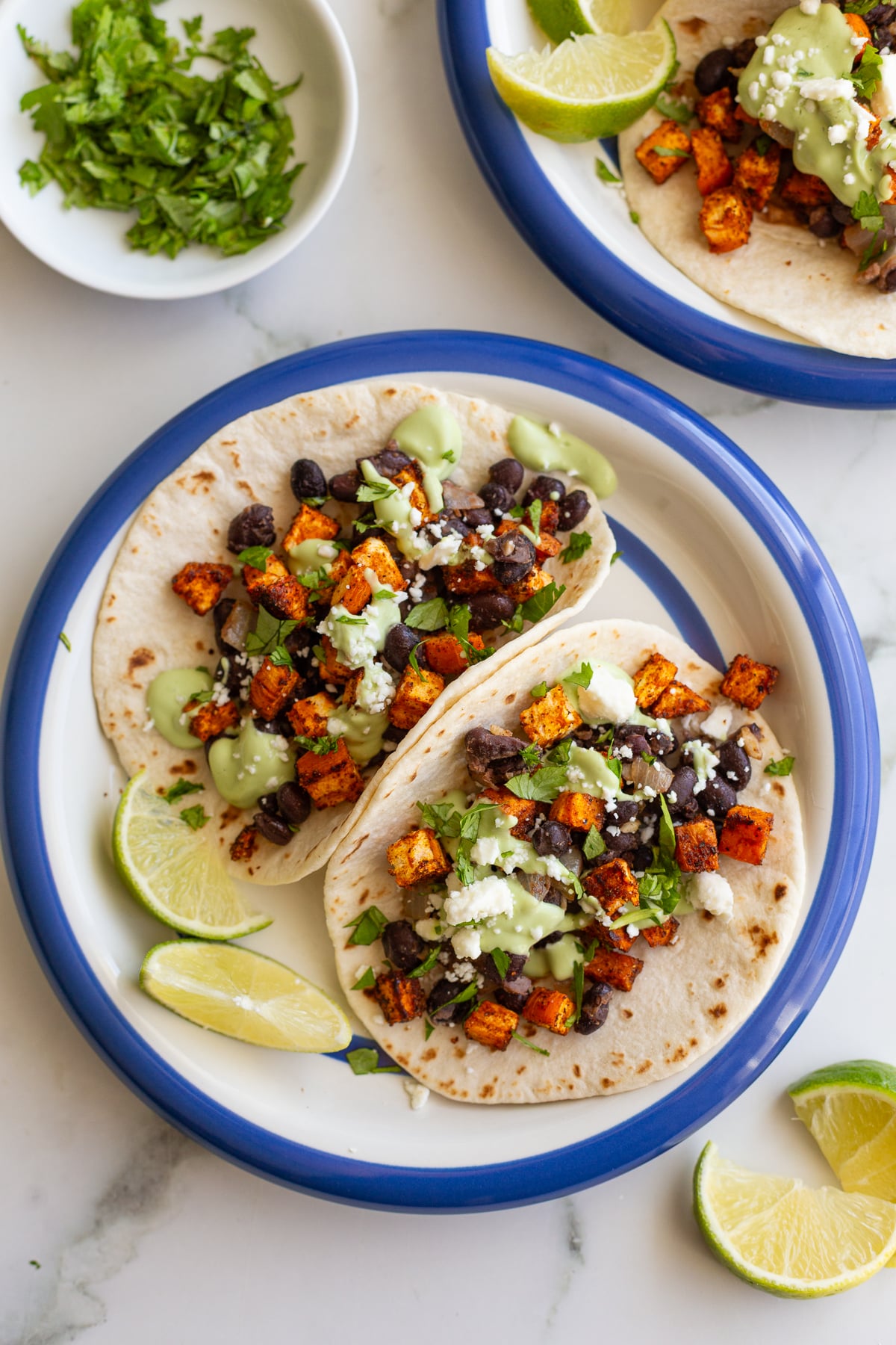 sweet potato and black bean tacos drizzled with an avocado crema. they're sitting on a white plate with royal blue rim