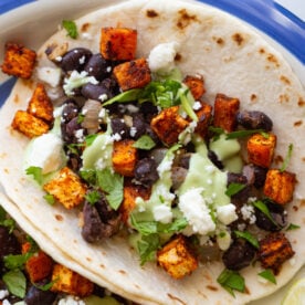 sweet potato and black bean tacos drizzled with an avocado crema. they're sitting on a white plate with royal blue rim