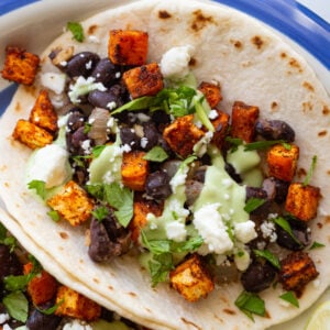 sweet potato and black bean tacos drizzled with an avocado crema. they're sitting on a white plate with royal blue rim