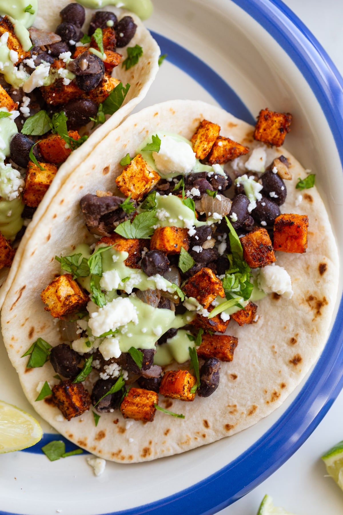 sweet potato and black bean tacos drizzled with an avocado crema. they're sitting on a white plate with royal blue rim