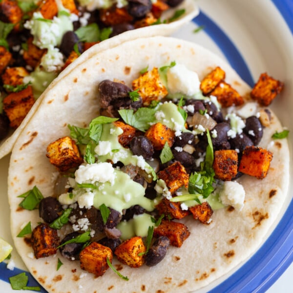 sweet potato and black bean tacos drizzled with an avocado crema. they're sitting on a white plate with royal blue rim