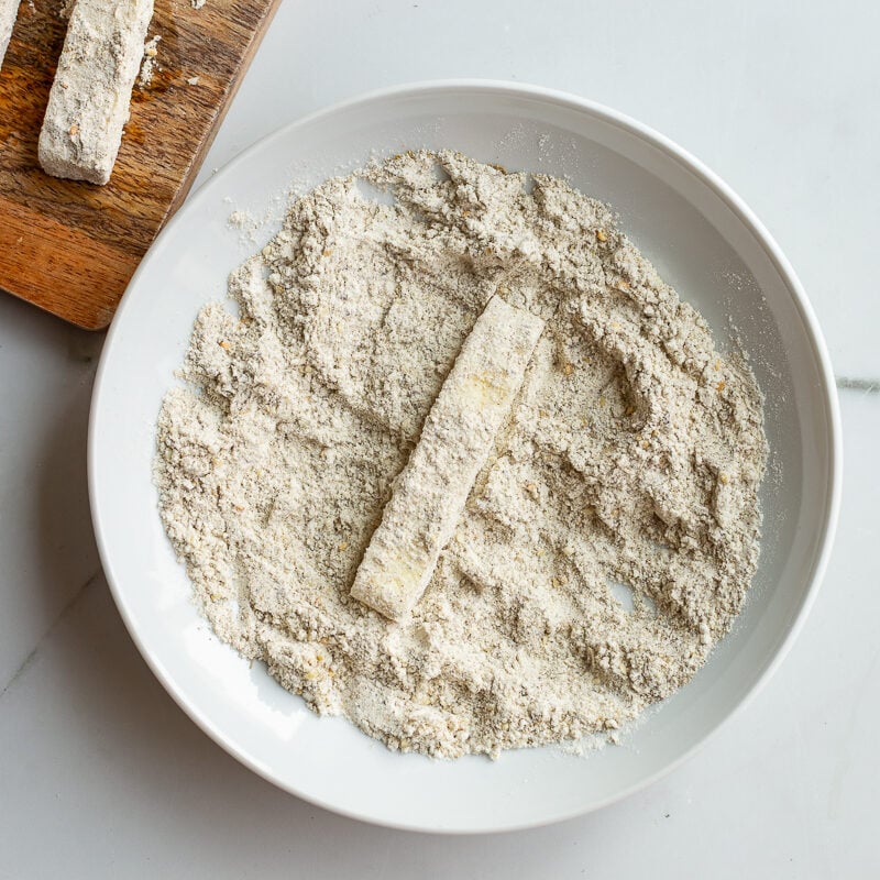 flour and zaatar mixed together in a shallow bowl with a piece of halloumi being dredged in the bowl