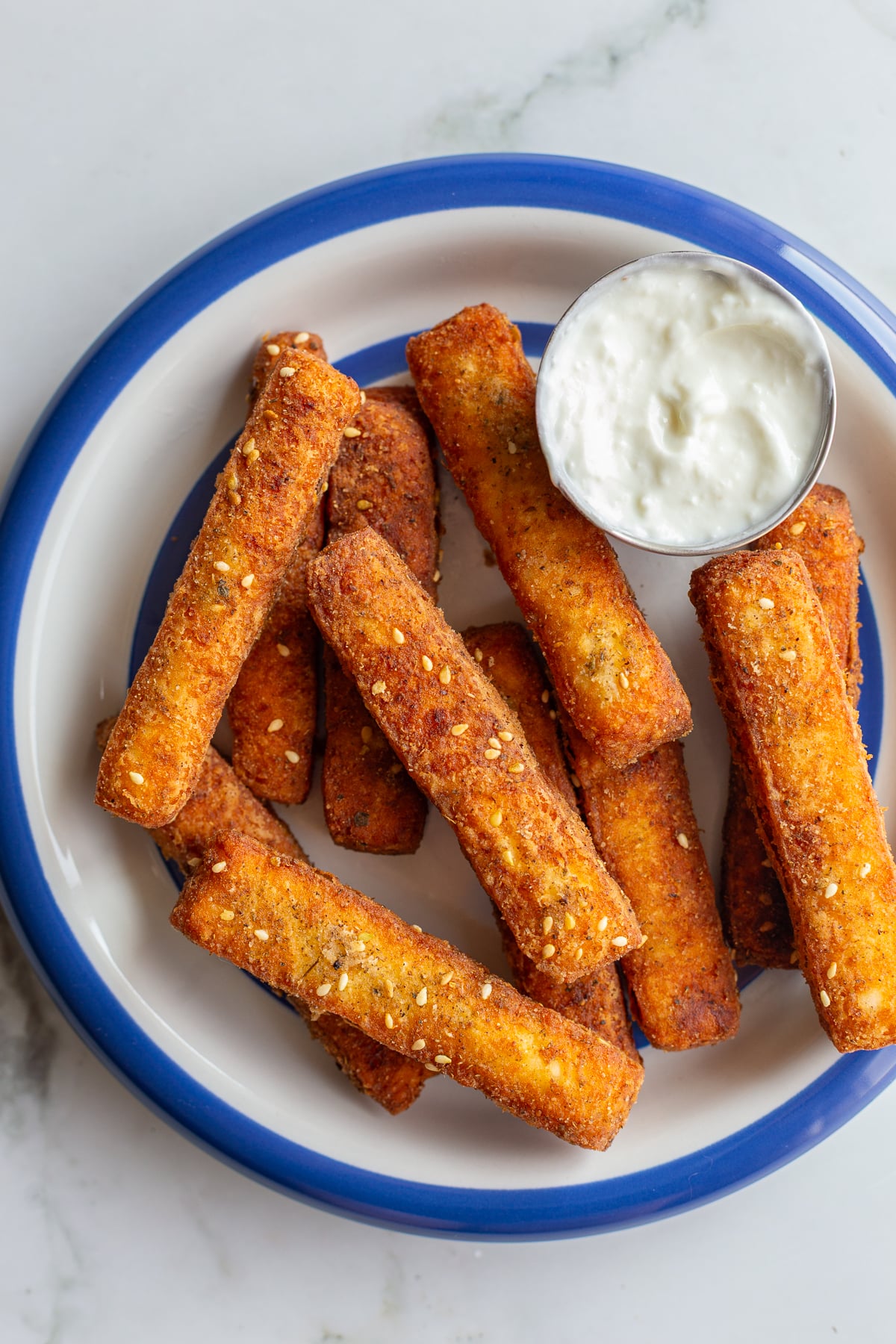 halloumi fries on a white plate with a blue rim. Yogurt sauce is in a small dish on the plate