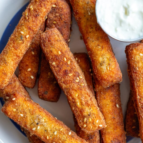 halloumi fries on a white plate with a blue rim. Yogurt sauce is in a small dish on the plate