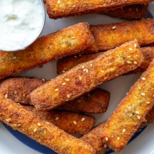 halloumi fries on a white plate with a blue rim. Yogurt sauce is in a small dish on the plate