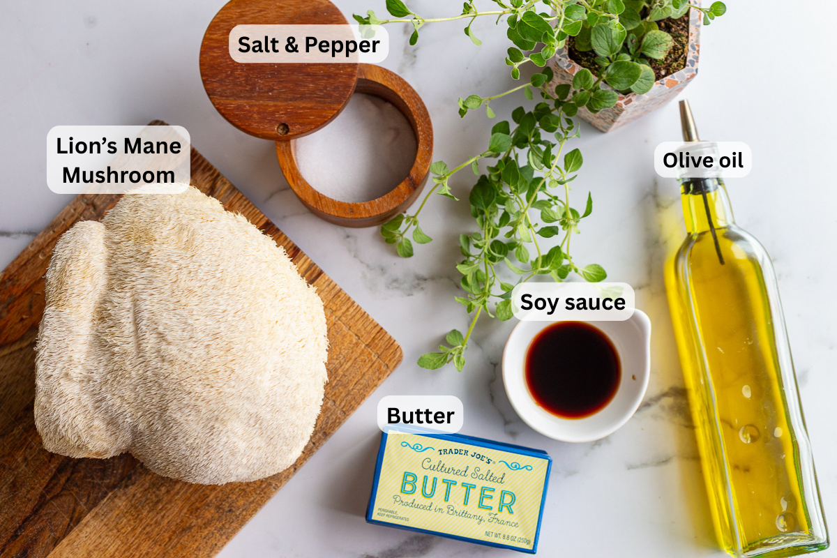 Overhead shot of lion's mane steak ingredients labeled on a marble counter, including lion's mane mushroom, butter, soy sauce, olive oil, salt and pepper, and fresh herbs
