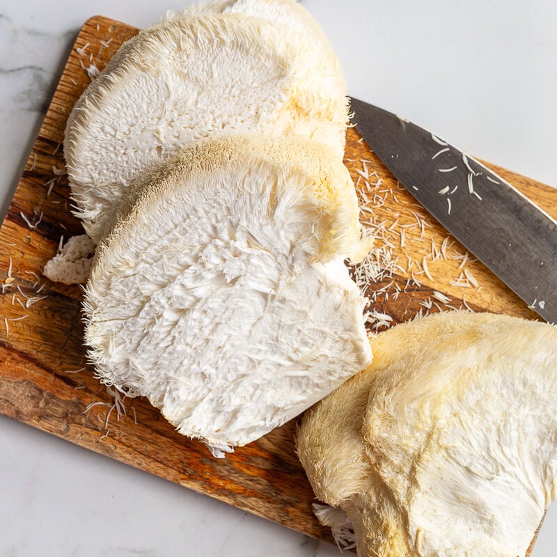 Overhead shot of lion's mane steak ingredients labeled on a marble counter, including lion's mane mushroom, butter, soy sauce, olive oil, salt and pepper, and fresh herbs
