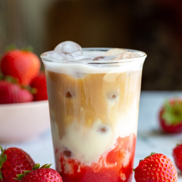 strawberry iced latte in a glass cup with fresh strawberries around the shot. You can clearly see the strawberry layer on the bottom with milk in the middle and a shot of espresso on the top