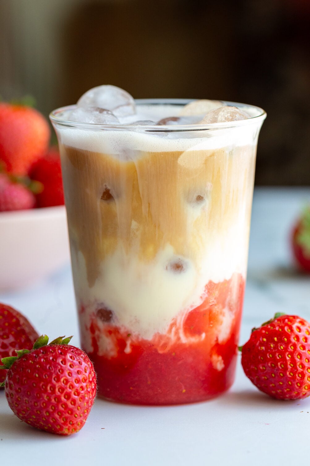 strawberry iced latte in a glass cup with fresh strawberries around the shot. You can clearly see the strawberry layer on the bottom with milk in the middle and a shot of espresso on the top