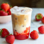 strawberry iced latte in a glass cup with fresh strawberries around the shot. You can clearly see the strawberry layer on the bottom with milk in the middle and a shot of espresso on the top