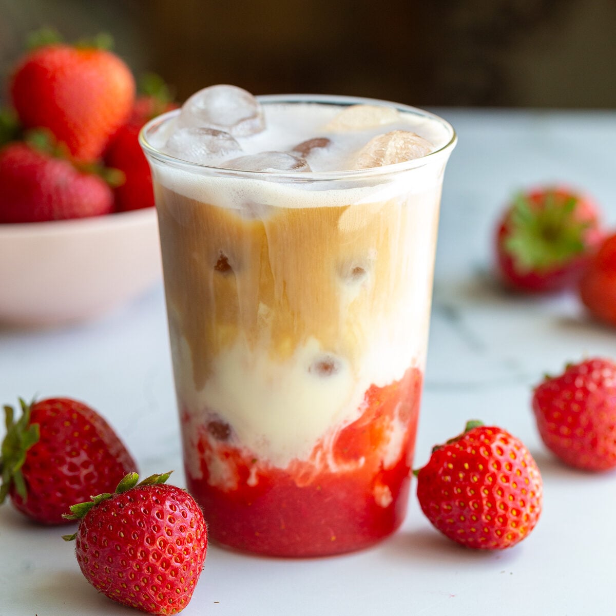 strawberry iced latte in a glass cup with fresh strawberries around the shot. You can clearly see the strawberry layer on the bottom with milk in the middle and a shot of espresso on the top