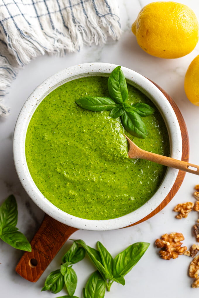 Overhead shot of walnut pesto in a white bowl on a wooden board, topped with a basil sprig and a wooden spoon, with lemons, fresh basil, and walnuts scattered around