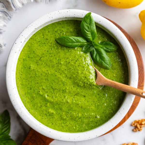 Walnut pesto in a white bowl on a wooden board with a wooden spoon resting inside, surrounded by fresh basil, lemons, and walnuts