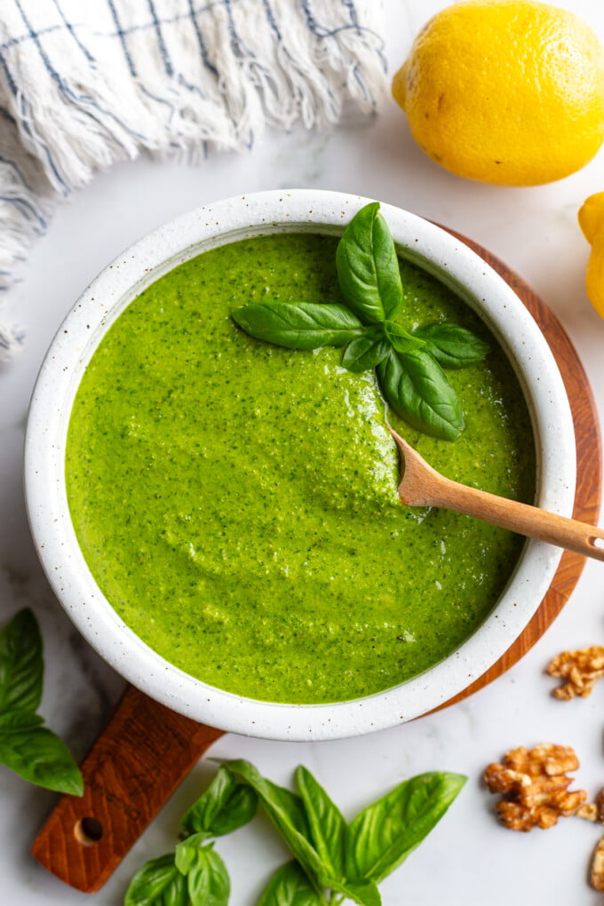 Walnut pesto in a white bowl on a wooden board with a wooden spoon resting inside, surrounded by fresh basil, lemons, and walnuts
