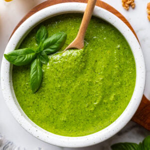 Walnut pesto in a white bowl on a wooden board with a wooden spoon resting inside, surrounded by fresh basil, lemons, and walnuts