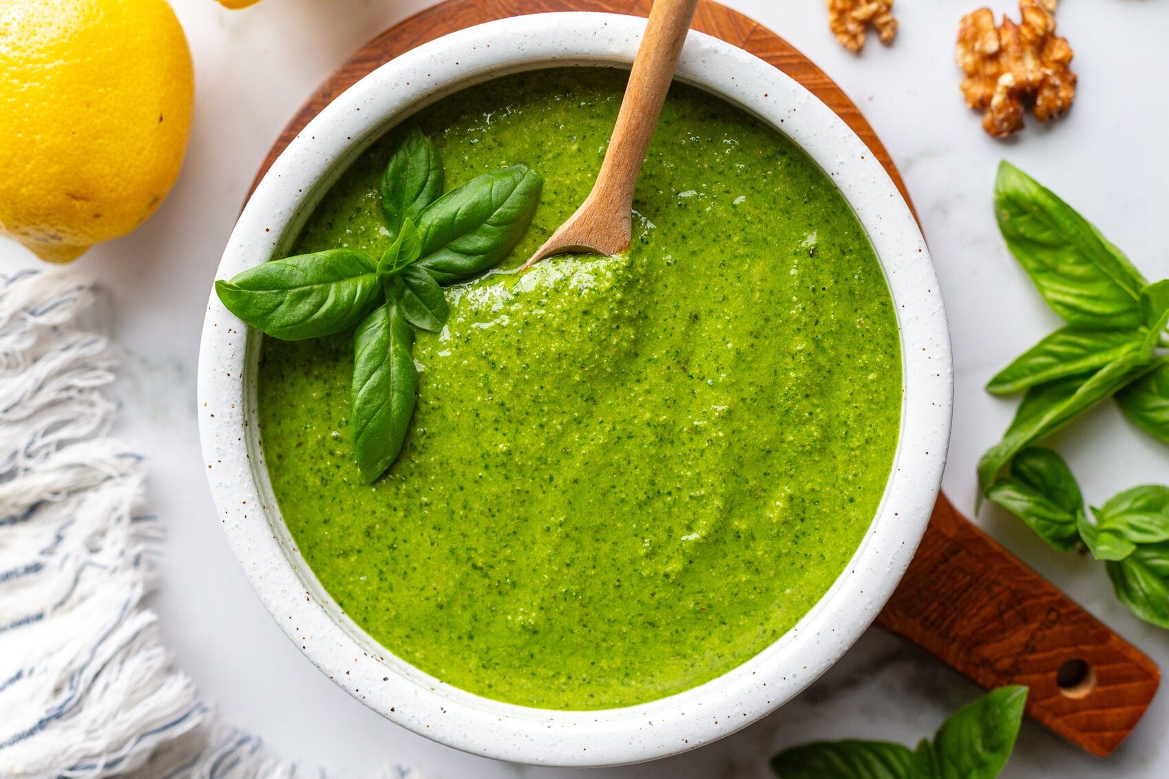 Walnut pesto in a white bowl on a wooden board with a wooden spoon resting inside, surrounded by fresh basil, lemons, and walnuts