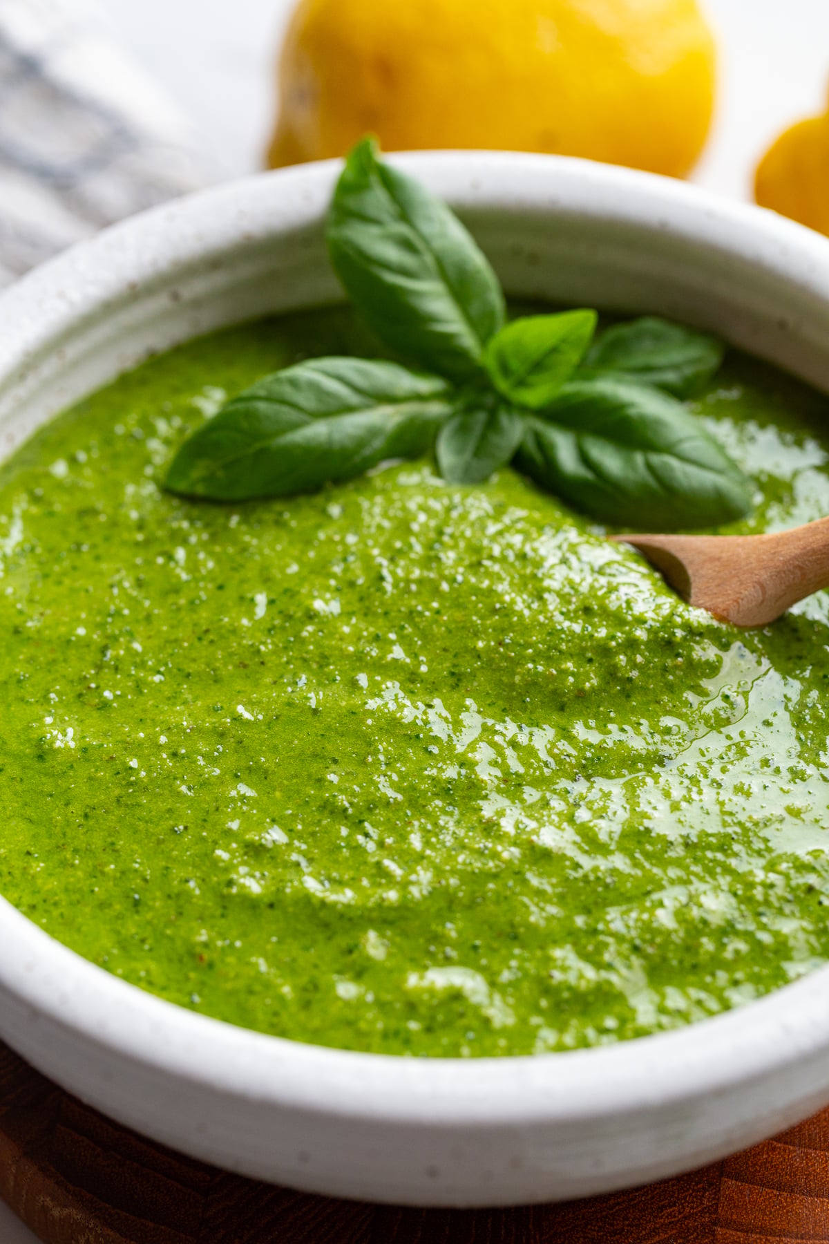 Close-up of walnut pesto in a white bowl garnished with basil, with lemons blurred in the background