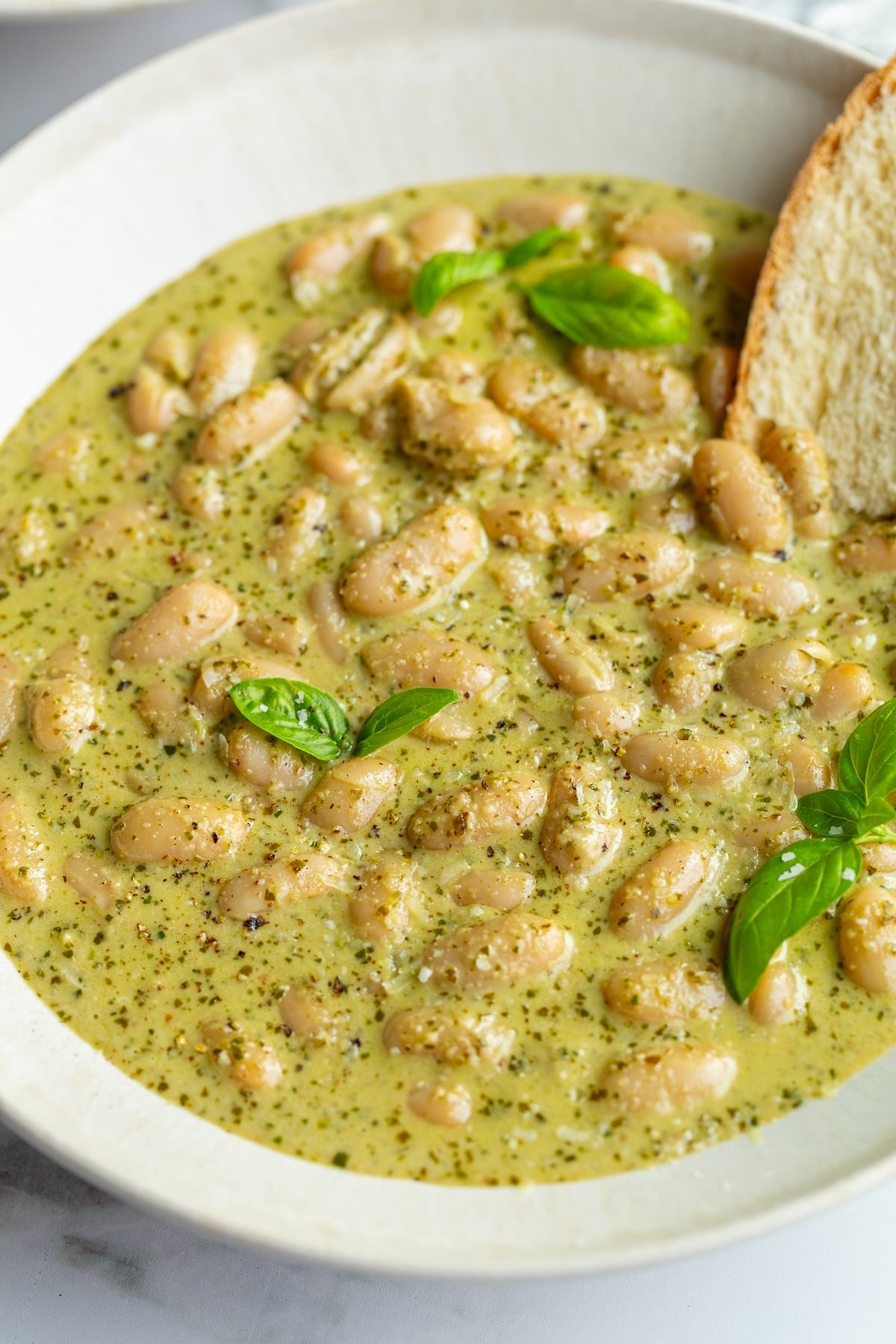 up close shot of big bowl of creamy pesto beans topped with fresh basil and fresh bread hanging out the side
