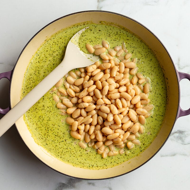 a creamy pesto sauce in a large skillet with cannellini beans poured on top before being mixed in