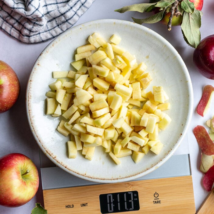 apple pieces on a plate on a kitchen scale with lots of apples and spple scrapes around the scene. towel in the corner and fresh apples around