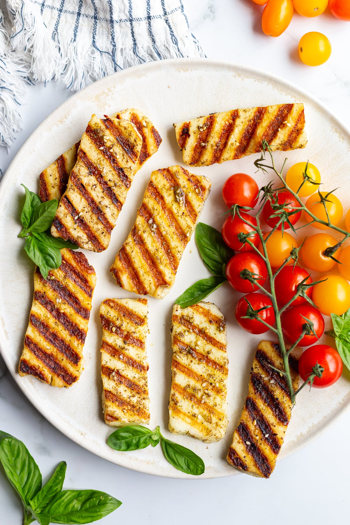 Overhead shot of grilled halloumi slices with charred grill marks on a white plate, garnished with fresh basil and served alongside cherry tomatoes on the vine
