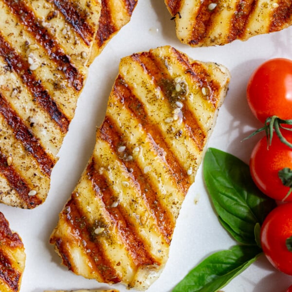 Close overhead shot of grilled halloumi with golden grill marks, seasoning, and cherry tomatoes on a white plate