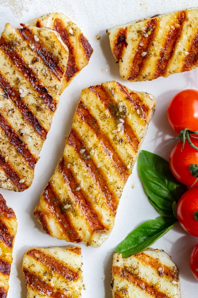 Close overhead shot of grilled halloumi with golden grill marks, seasoning, and cherry tomatoes on a white plate