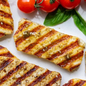 Close overhead shot of grilled halloumi with golden grill marks, seasoning, and cherry tomatoes on a white plate