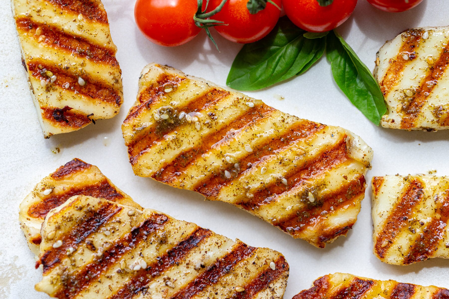 Close overhead shot of grilled halloumi with golden grill marks, seasoning, and cherry tomatoes on a white plate