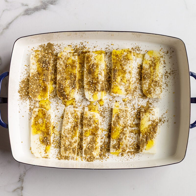 Sliced halloumi in a baking dish drizzled with olive oil and sprinkled with zaatar seasoning