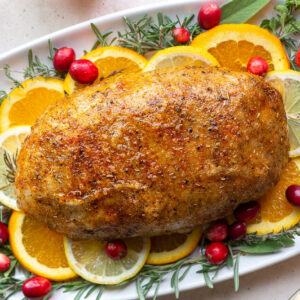 vegan turkey loaf sitting on a bed of fresh herbs and citrus slices. plates of green beans, pumpkin pie, and mashed potatoes are in the corner of the shot