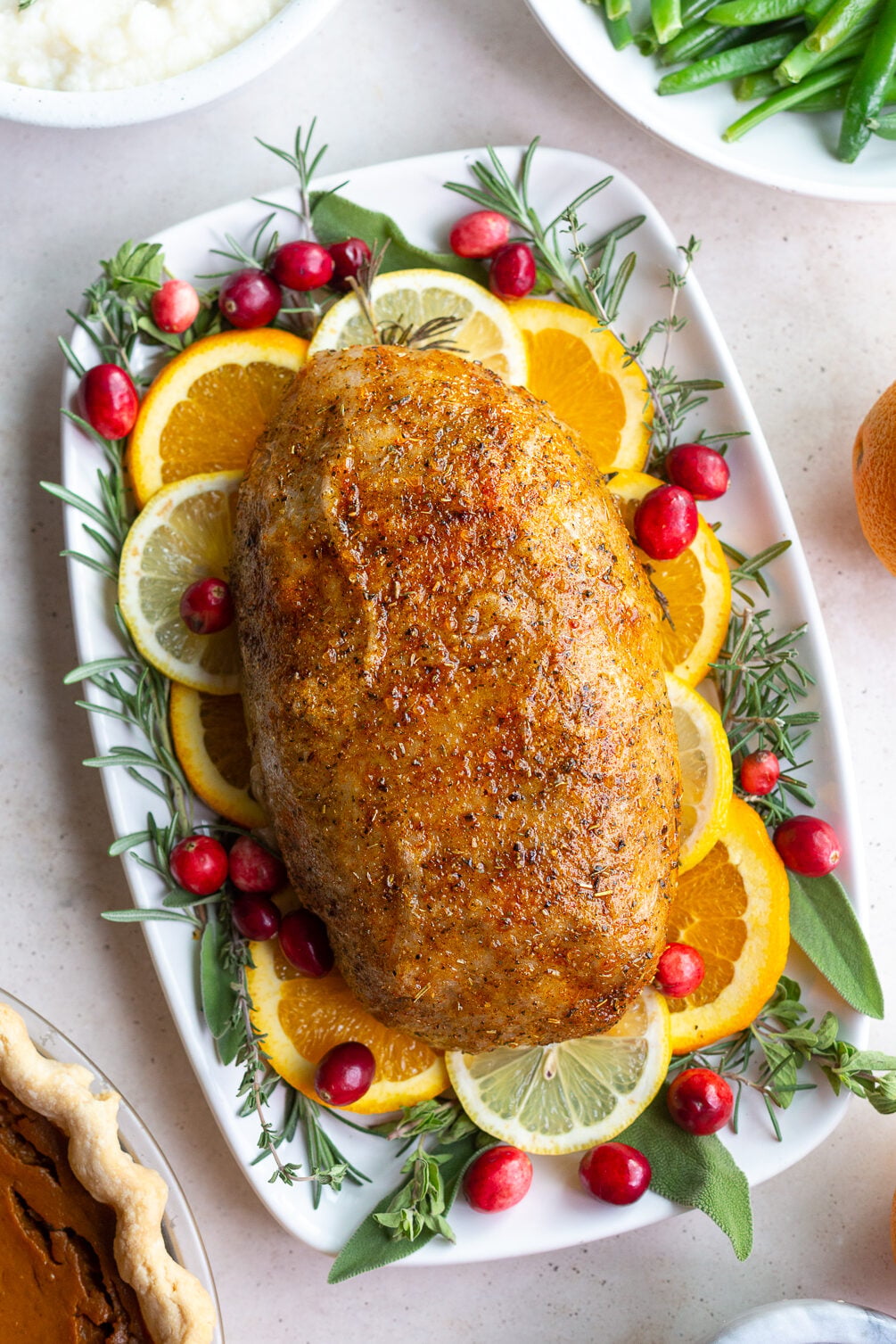vegan turkey loaf sitting on a bed of fresh herbs and citrus slices. plates of green beans, pumpkin pie, and mashed potatoes are in the corner of the shot